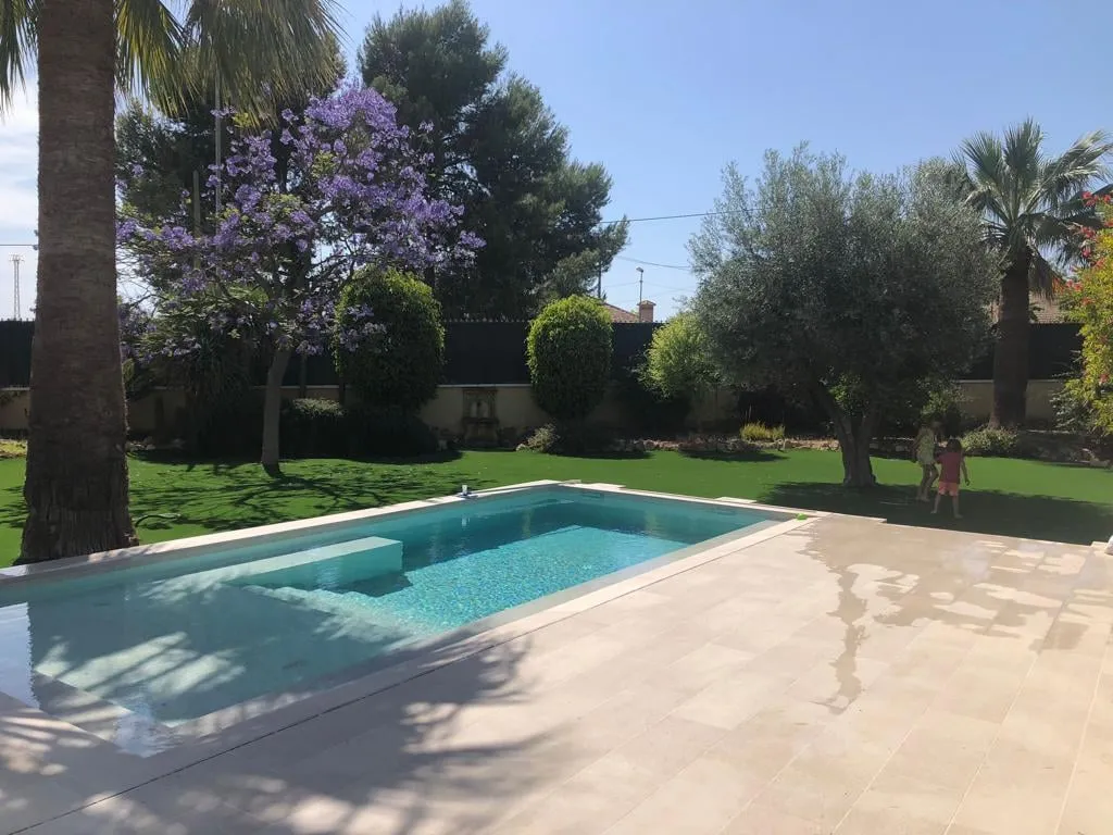 Piscina con borde de piedra clara y jardín con césped, olivos y árboles en flor
